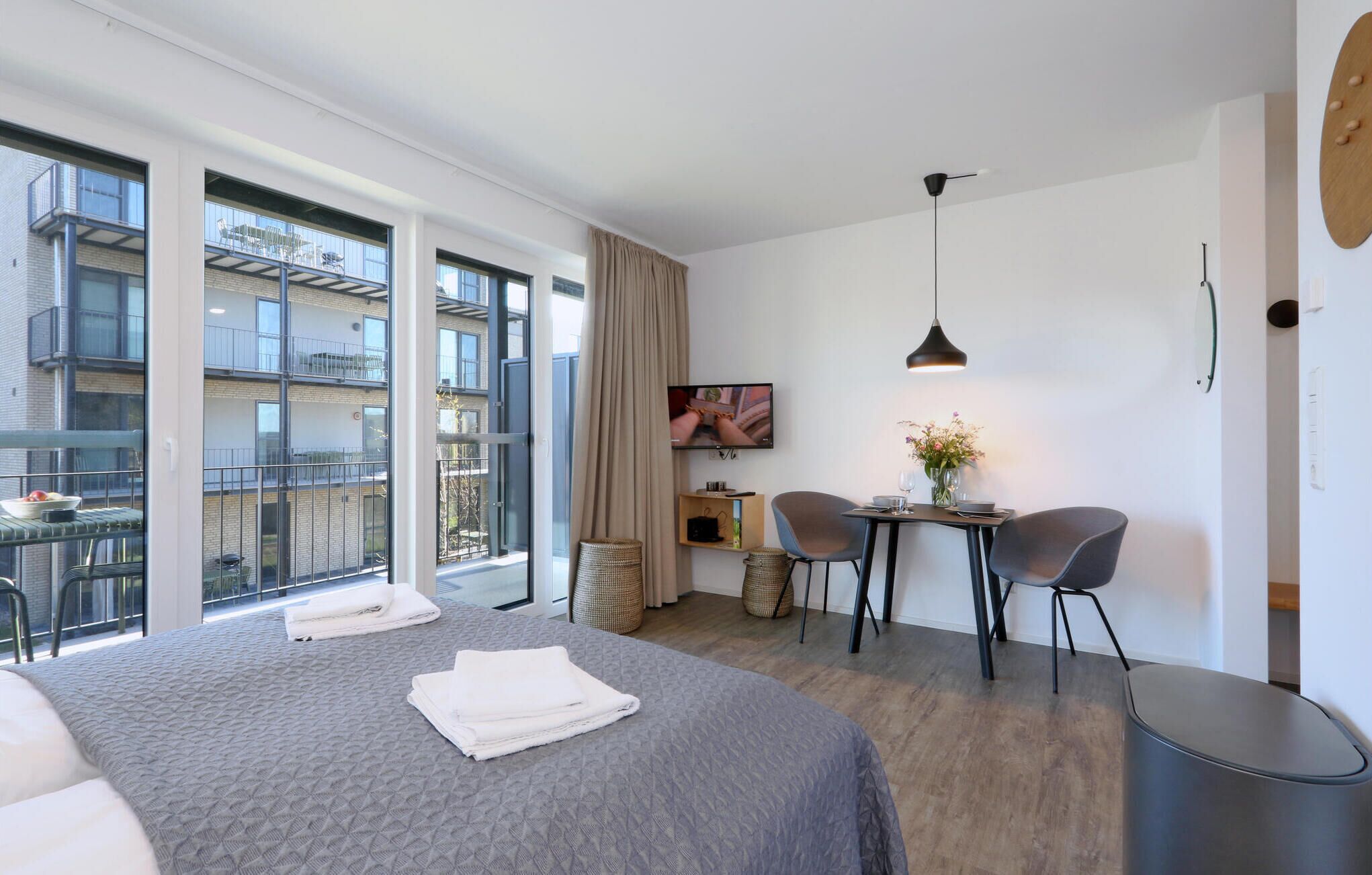 A modern hotel room with a large bed, a desk with two chairs, a TV, and a balcony with a view of other buildings.