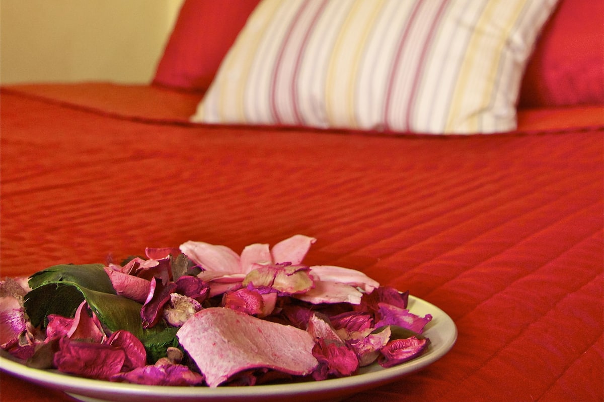 A bed with a red bedspread, a plate of dried flowers in the foreground, and a wall-mounted shelf with a clock.