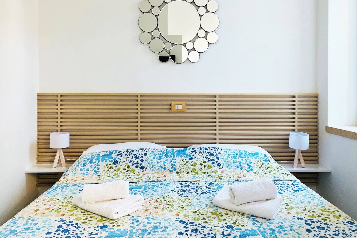 A neatly made bed with a floral-patterned bedspread, two bedside lamps, a wooden headboard, and a wall-mounted mirror.