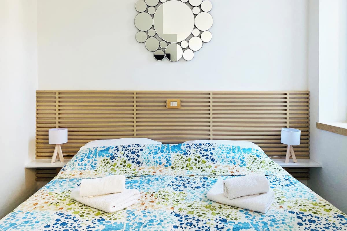 A neatly made bed with a floral-patterned bedspread, two bedside lamps, a wooden headboard, and a wall-mounted mirror.