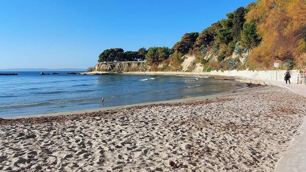 A beach with a curved shoreline, a stone wall, and a person walking along the path. - En-suite Iris on Rokova Street (Split)