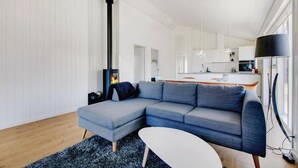 A modern living room with a grey sectional sofa, a small white table, and a black floor lamp. A black fireplace with a visible fire is in the corner. The room has white walls and a wooden floor. - Nice home in Glesborg with sauna (Glesborg)