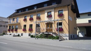 A two-story building with a yellow facade, multiple windows with flower boxes, and a visible entrance with steps. - Gasthof Bauer (Elsendorf)