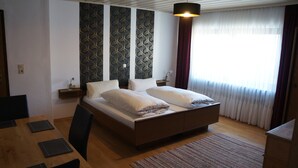A bedroom with a wooden bed, a dining table with chairs, a patterned wall, and a window with curtains. - Gasthof Bauer (Elsendorf)