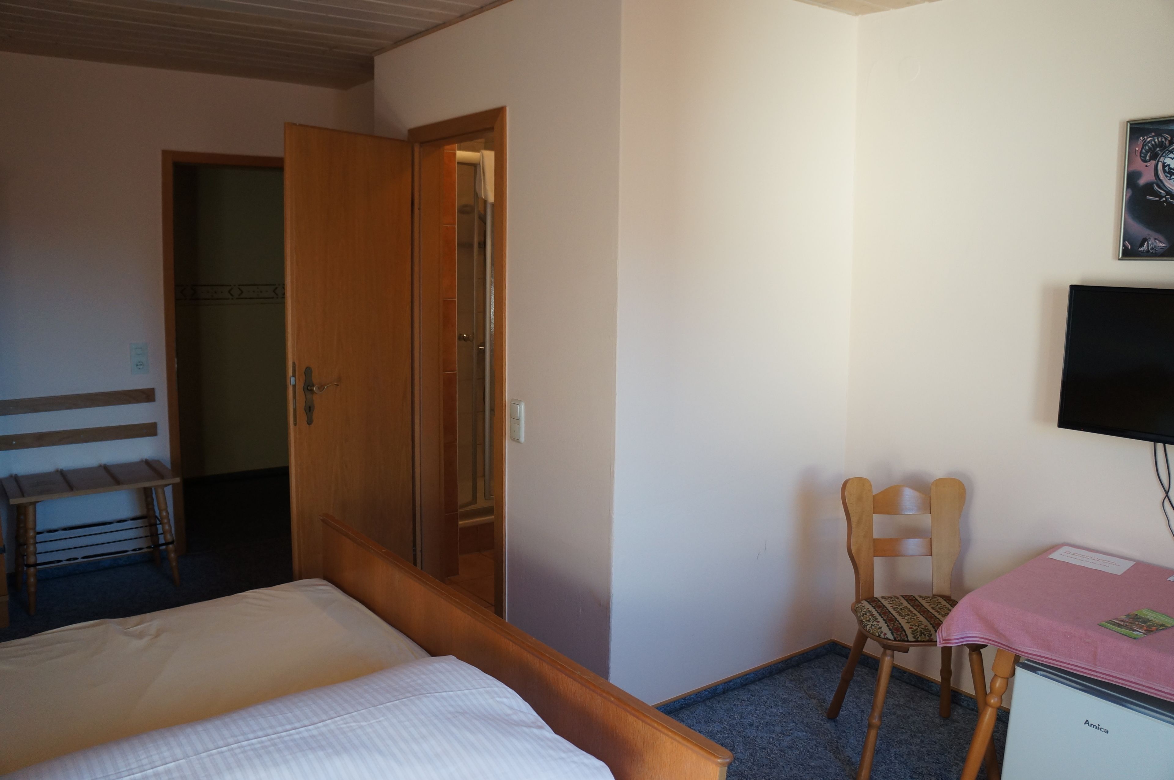 Habitación de hotel con cama, una silla de madera, un escritorio con mantel rosa, televisor y armario.