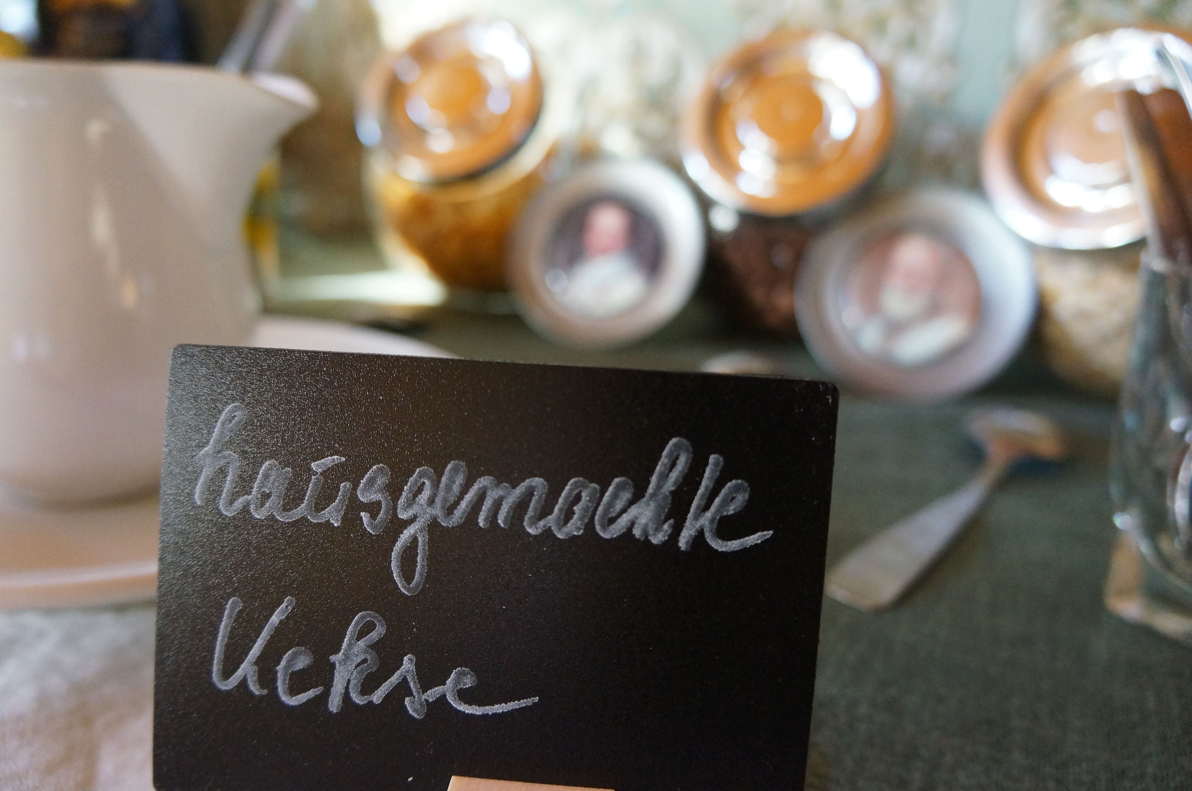 Un gros plan d’une pancarte noire avec du texte manuscrit dans une langue étrangère, une tasse blanche et des bocaux avec des couvercles en arrière-plan.