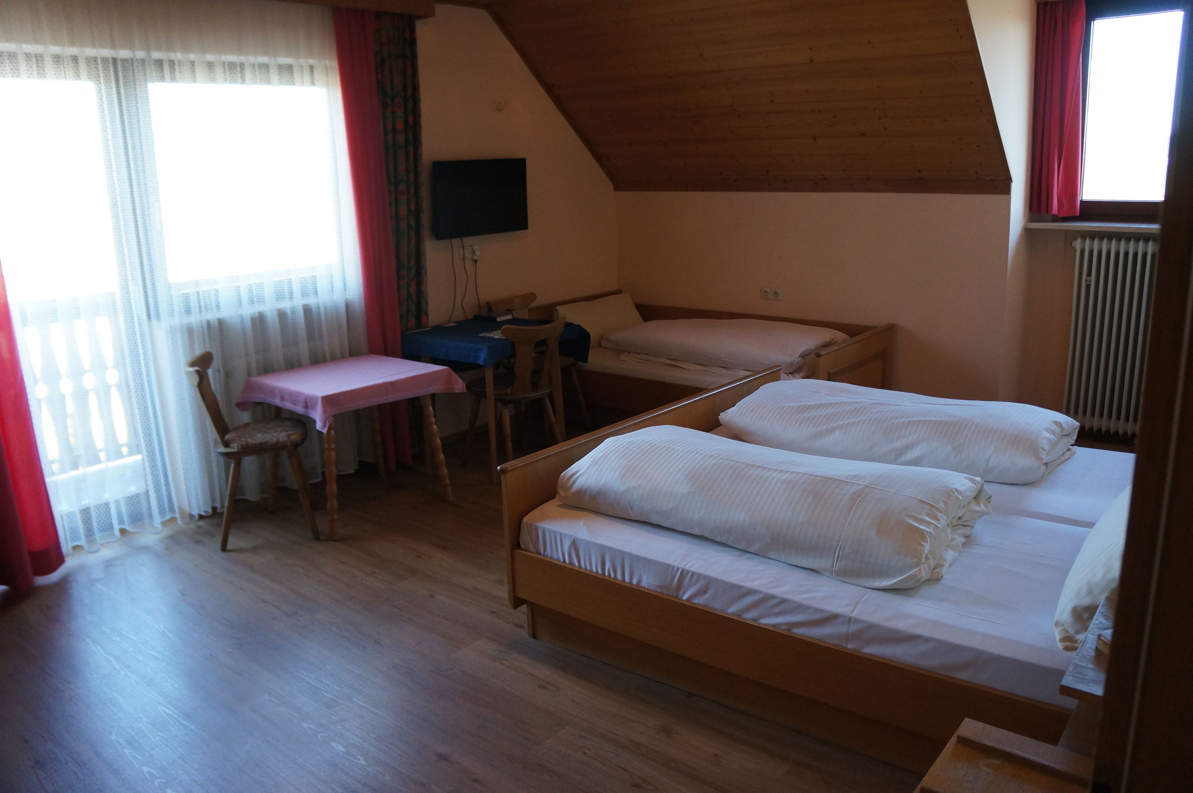 A bedroom with a wooden ceiling, a desk with a chair, a bed with two pillows, and a TV on the wall.