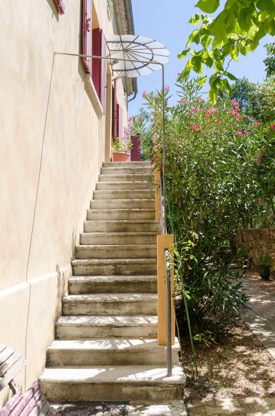 Eine Treppe aus Stein führt zu einem Gebäude mit einem roten Fensterlädchen und einer weißen, umhüllenden Struktur auf der linken Seite.