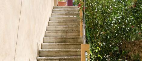 Eine Treppe aus Stein führt zu einem Gebäude mit einem roten Fensterlädchen und einer weißen, umhüllenden Struktur auf der linken Seite.