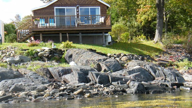 A cabin with a deck, large windows, and a view of a rocky shoreline and water.