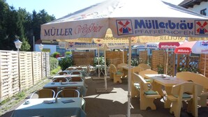 An outdoor seating area with tables and chairs under a canopy with 'Müller Weisbier' and 'Müllerbräu' signage.