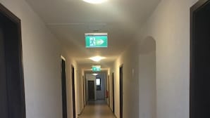 A long, narrow hallway with a light-colored floor and white walls. There are doors on both sides of the hallway, and a green emergency exit sign is visible on the ceiling.