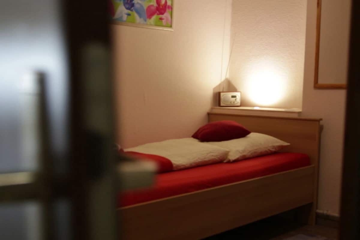 Un letto singolo con materasso rosso e biancheria da letto bianca, un comodino con una lampada e un quadro floreale al muro.