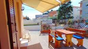 A covered outdoor seating area with a table and benches, a door, and a view of the street and buildings. - N044 - Numana, new three-room apartment with terrace and a / c (Numana)