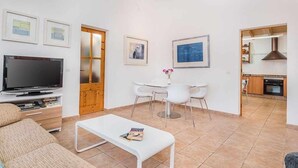 Een moderne woonkamer met een flatscreen-tv, een witte salontafel en een bank. Er is een eethoek met een witte tafel en stoelen, en een keuken is zichtbaar door een open deur.