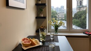 Une pièce avec une table décorée de fruits d’agrumes et un vase rempli de fleurs, une fenêtre donnant sur un paysage urbain et des étagères ornées de plantes.