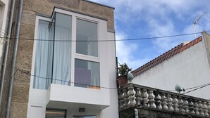 Un edificio de dos pisos con fachada blanca, un balcón con macetas y un muro de piedra adyacente.