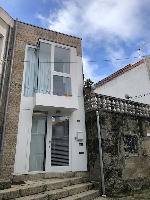 A two-story building with a white facade, a balcony with potted plants, and a stone wall adjacent to it.