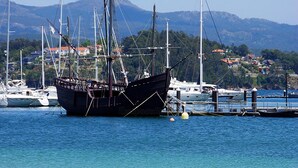 Una playa con un velero grande amarrado en un muelle, un cielo azul despejado y un paisaje montañoso al fondo.