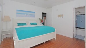 A bedroom with a white bed, a teal bedspread, a white nightstand, a lamp, and a painting on the wall. - The South Beach Party House (Miami Beach)