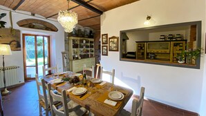 Un comedor con una mesa de madera dispuesta para cuatro personas, rodeada de sillas blancas. Un candelabro cuelga del techo, y se ve una estantería llena de libros y objetos decorativos al fondo.