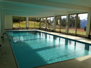 An indoor swimming pool with clear glass walls, a tiled floor, and a view of a grassy area and mountains outside. - Cransalpin SKI IN SKI OFF (Crans-Montana)