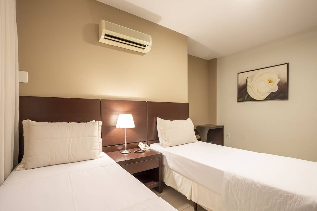 Habitación de hotel con dos camas, una mesita de noche con lámpara y teléfono, aire acondicionado y un cuadro de una flor en la pared.