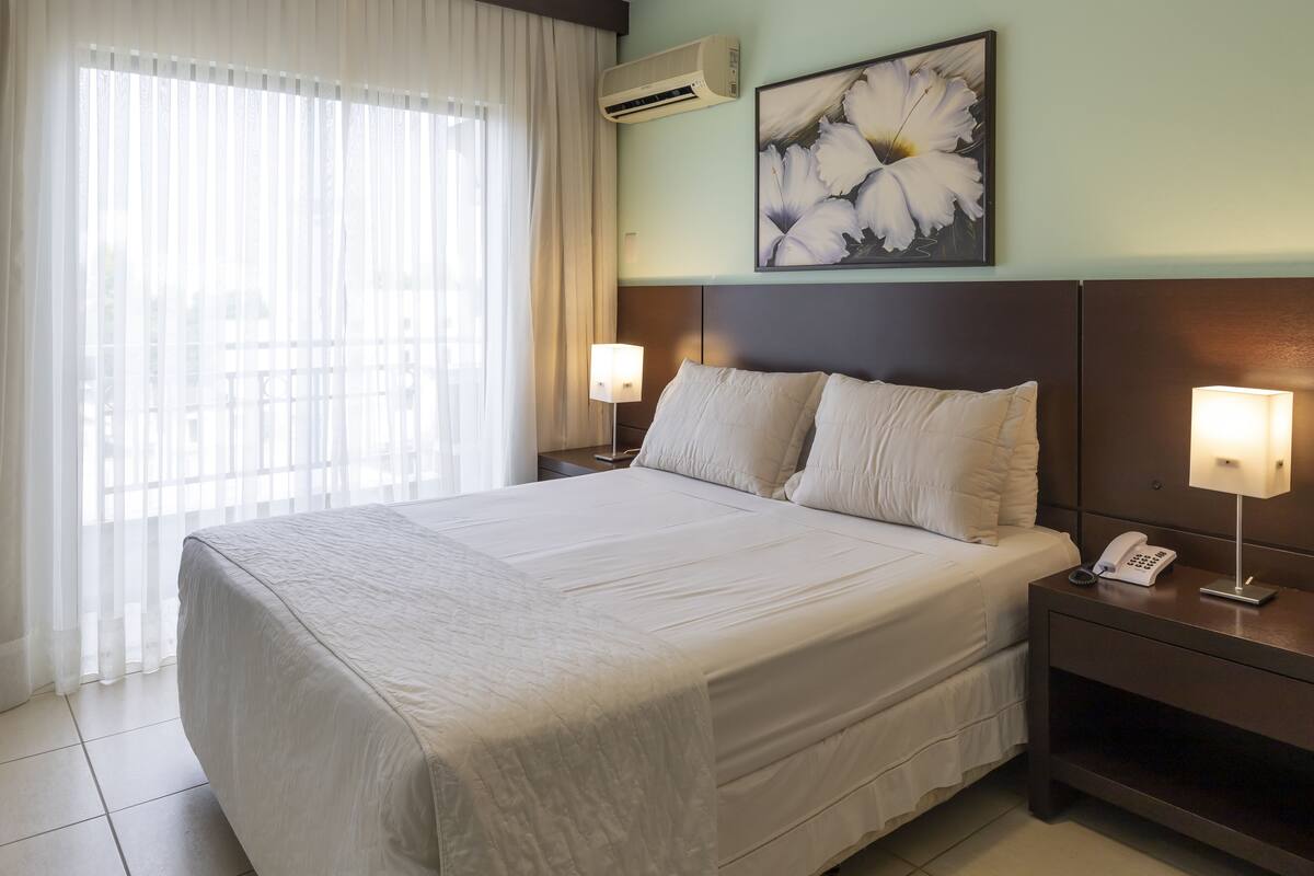 Habitación de hotel con una cama grande, dos mesitas de noche con lámparas, aire acondicionado de pared y un cuadro floral sobre la cama.