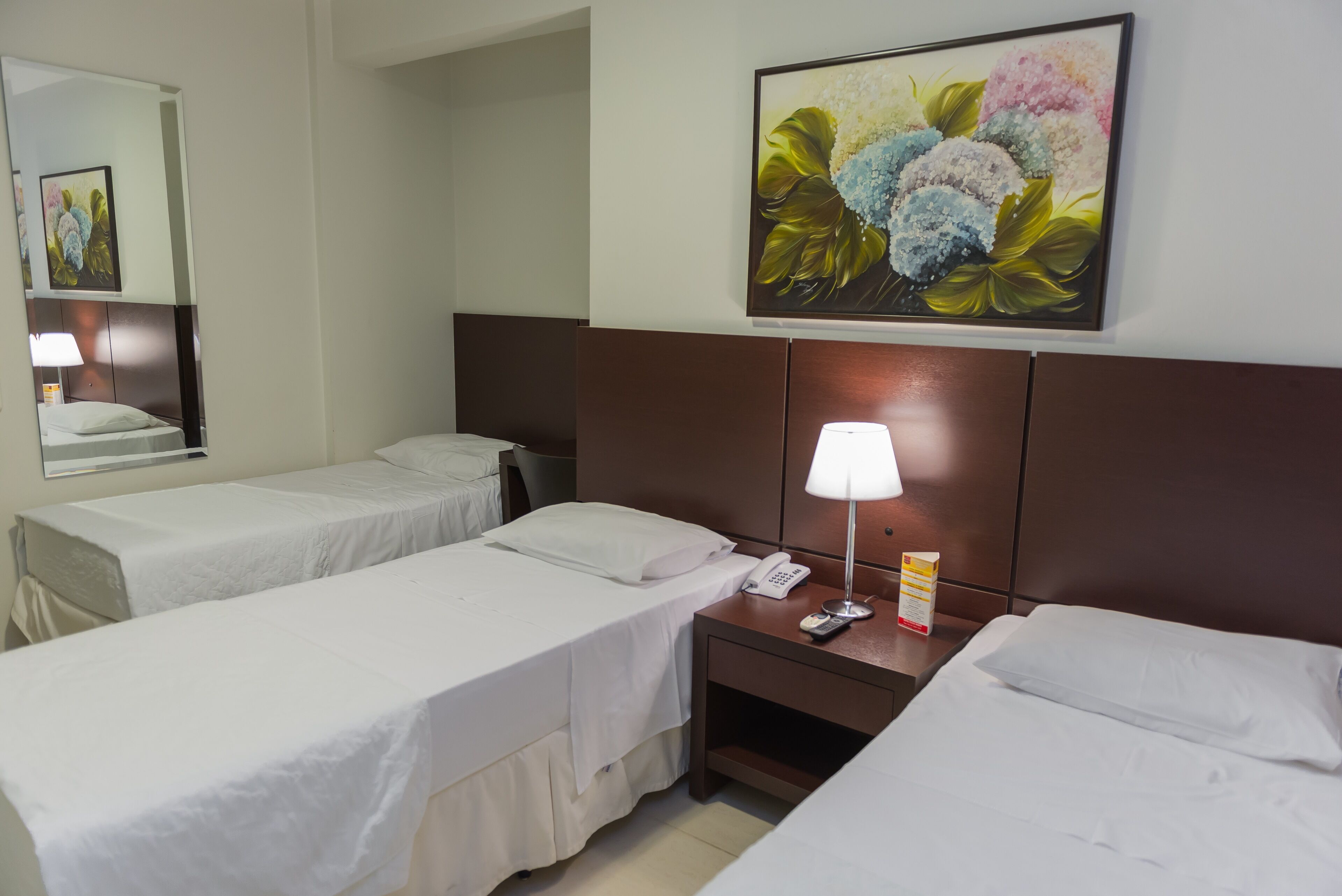 Habitación de hotel con dos camas individuales, una mesita de noche con lámpara, teléfono y un cuadro de flores en la pared.