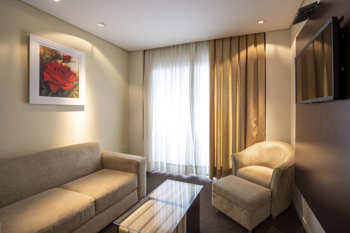 Una sala moderna con un sofá beige, una mesa de centro de vidrio y un sillón beige. Se ve un ventanal con cortinas translúcidas y vistas al exterior.