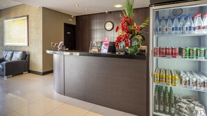 A hotel reception area with a counter, a clock, a refrigerator stocked with beverages, and a floral arrangement. - Hotel Plaza Norte (Joinville)