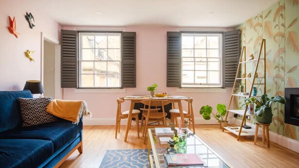 Una sala de estar con un sofá azul, una mesa redonda de madera y una mesita de centro. Hay dos ventanas con contraventanas, una estantería y una planta.