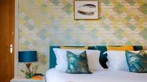 Una habitación de hotel con pared estampada, cama con manta amarilla, almohadas azul verdosas y blancas, y mesita de noche con lámpara y decoración de piña.