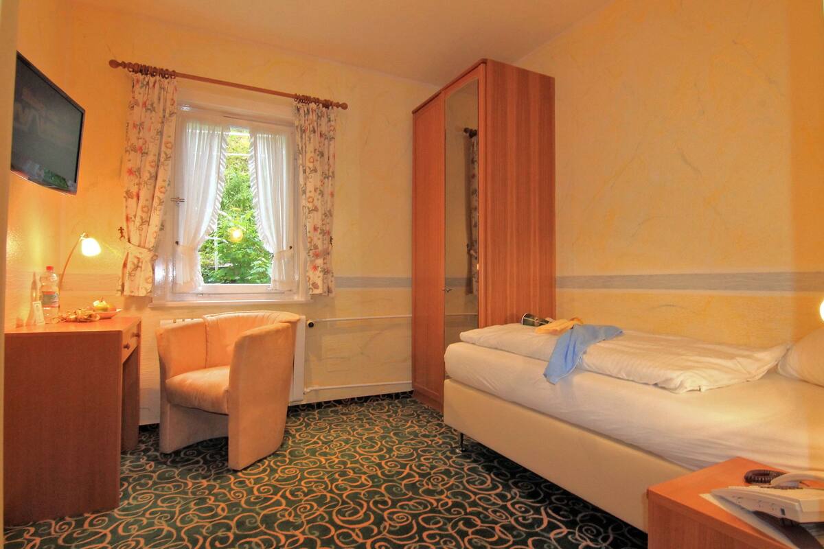 A hotel room with a bed, a chair, a desk with a lamp, a window with curtains, and a mirror.