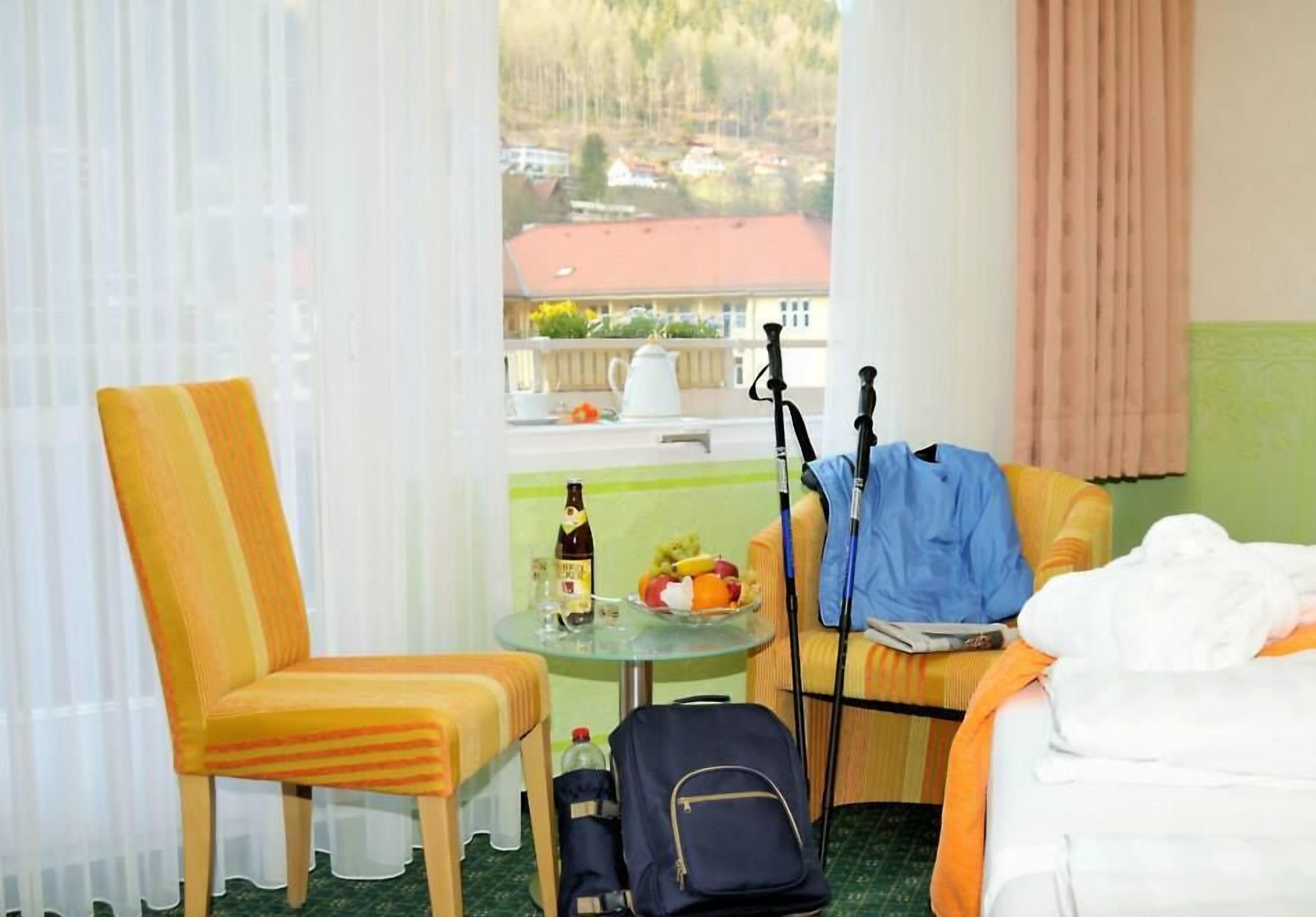 A hotel room with a bed, a chair, a table with a bottle and fruit, and a view of the outside.