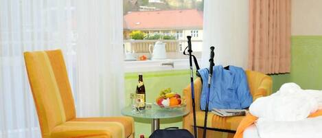 A hotel room with a bed, a chair, a table with a bottle and fruit, and a view of the outside.