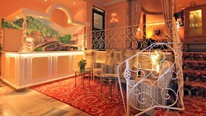 A hotel lobby with a reception desk, a mural of a bridge, a staircase with ornate railings, and a seating area with patterned upholstery.