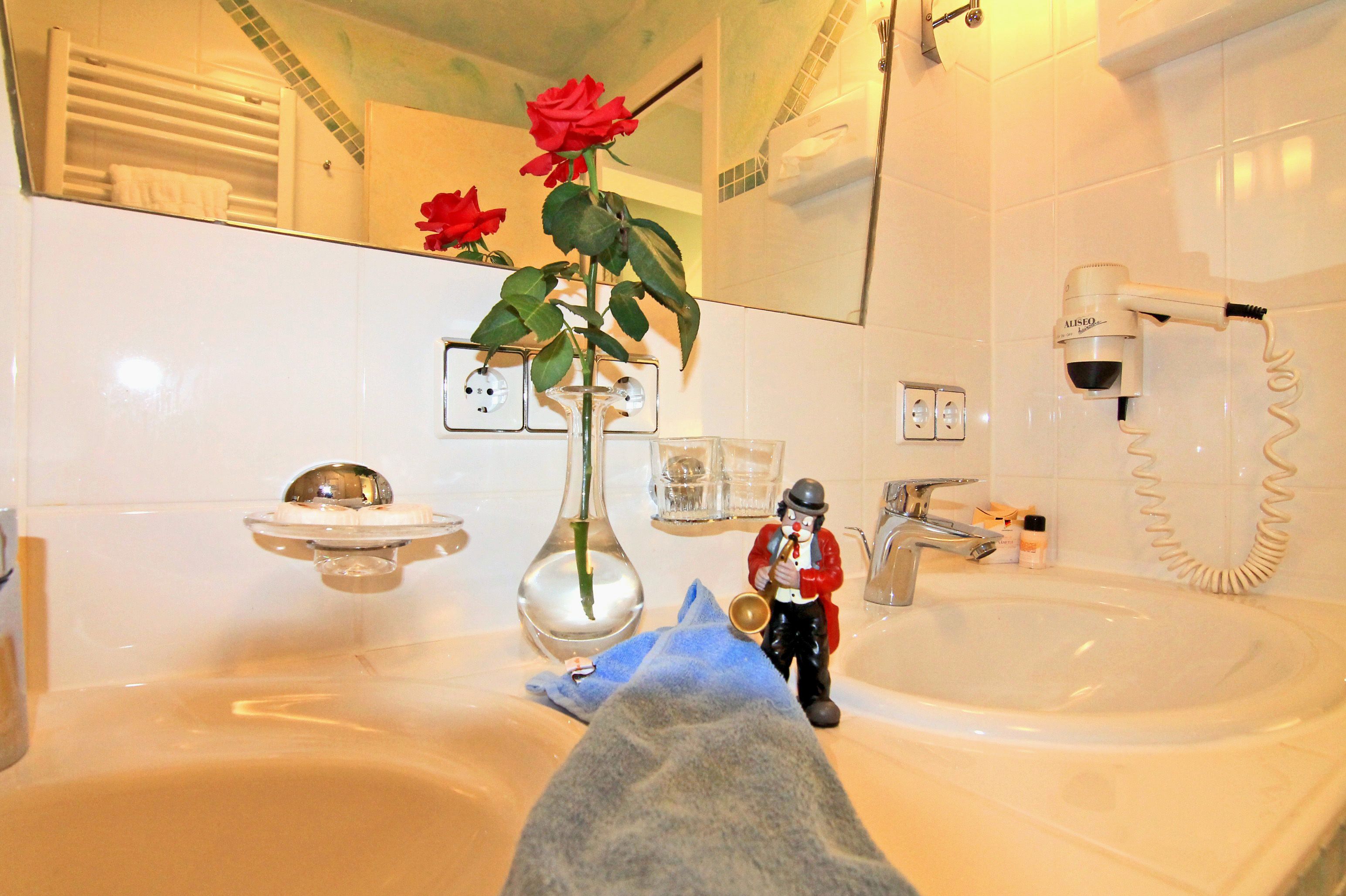 Un baño con lavamanos, espejo, toallero y una rosa roja en un jarrón.