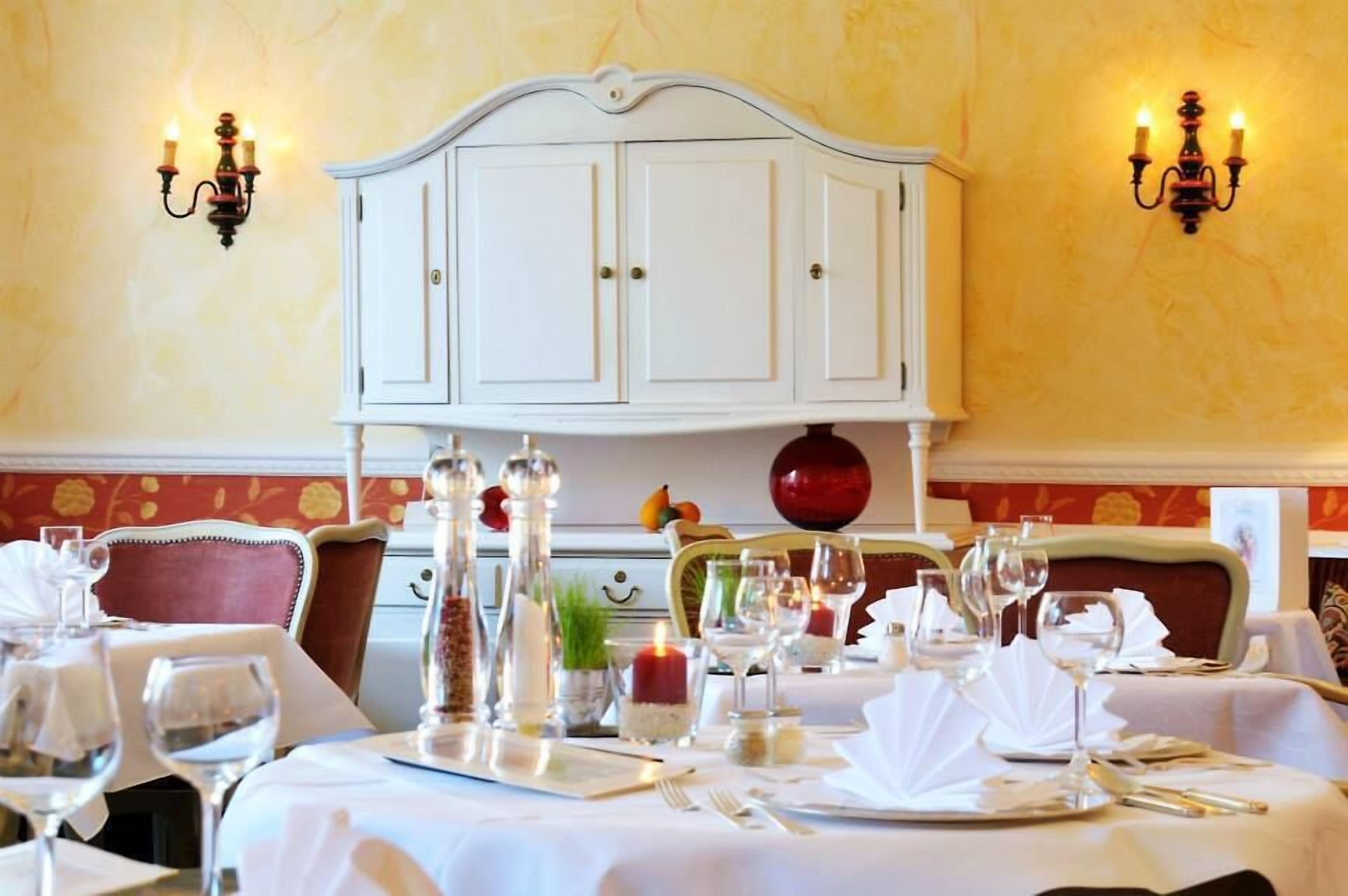 A dining table set with white tablecloths, candles, and wine glasses, with a white cabinet and wall sconces in the background.