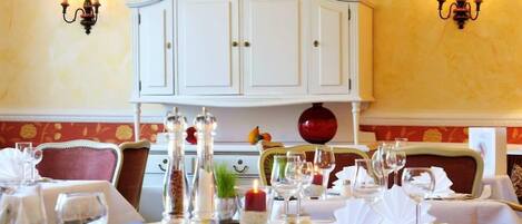 Una mesa de comedor dispuesta con manteles blancos, velas y copas de vino, con un aparador blanco y apliques de pared al fondo.
