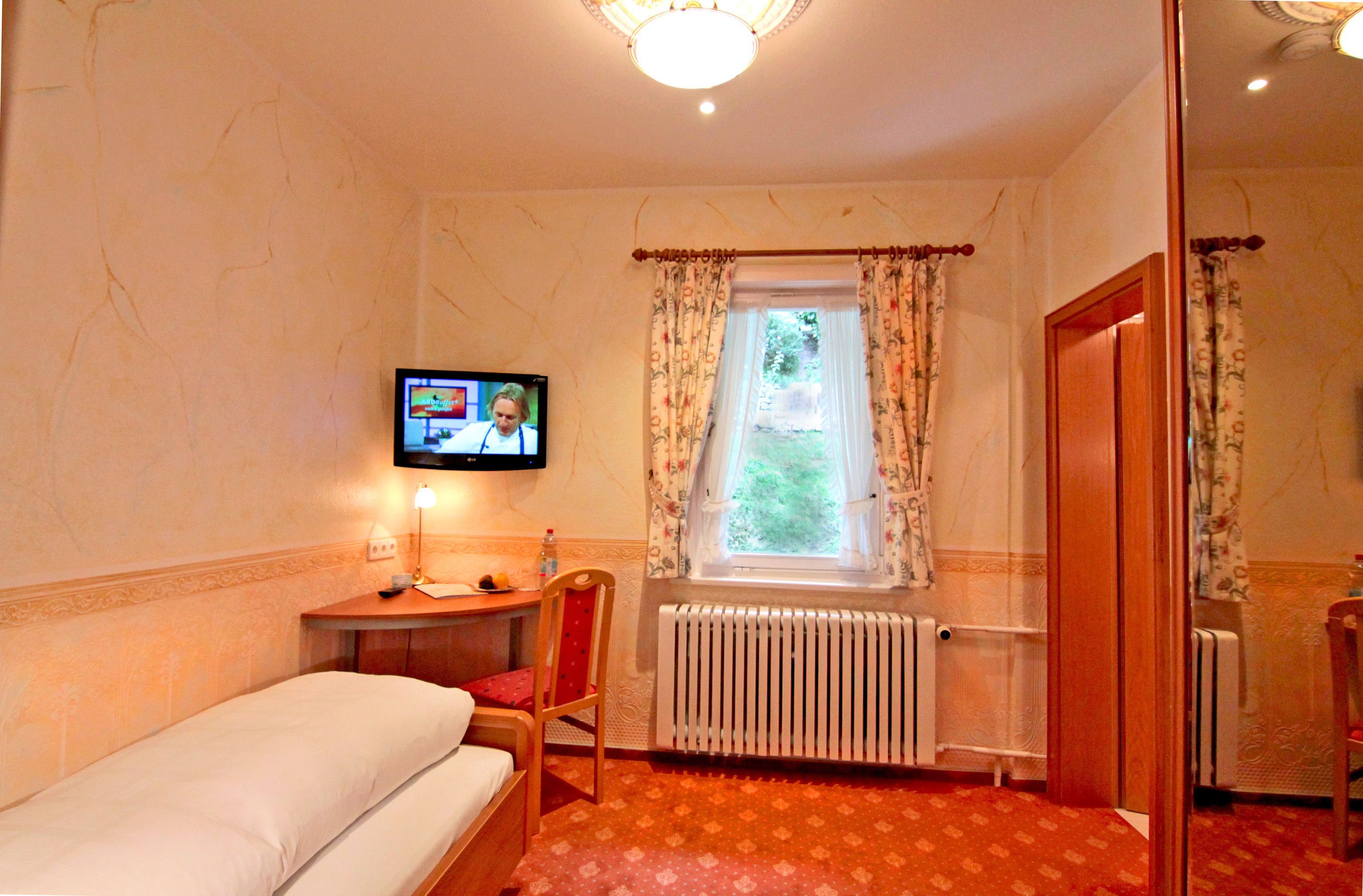 Una habitación de hotel con una cama individual, una mesita con una silla, un televisor montado en la pared, una ventana con cortinas y un radiador.
