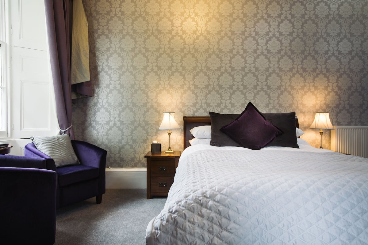 Une chambre à coucher avec un lit, deux tables de chevet avec des lampes, une chaise et une fenêtre avec des rideaux.