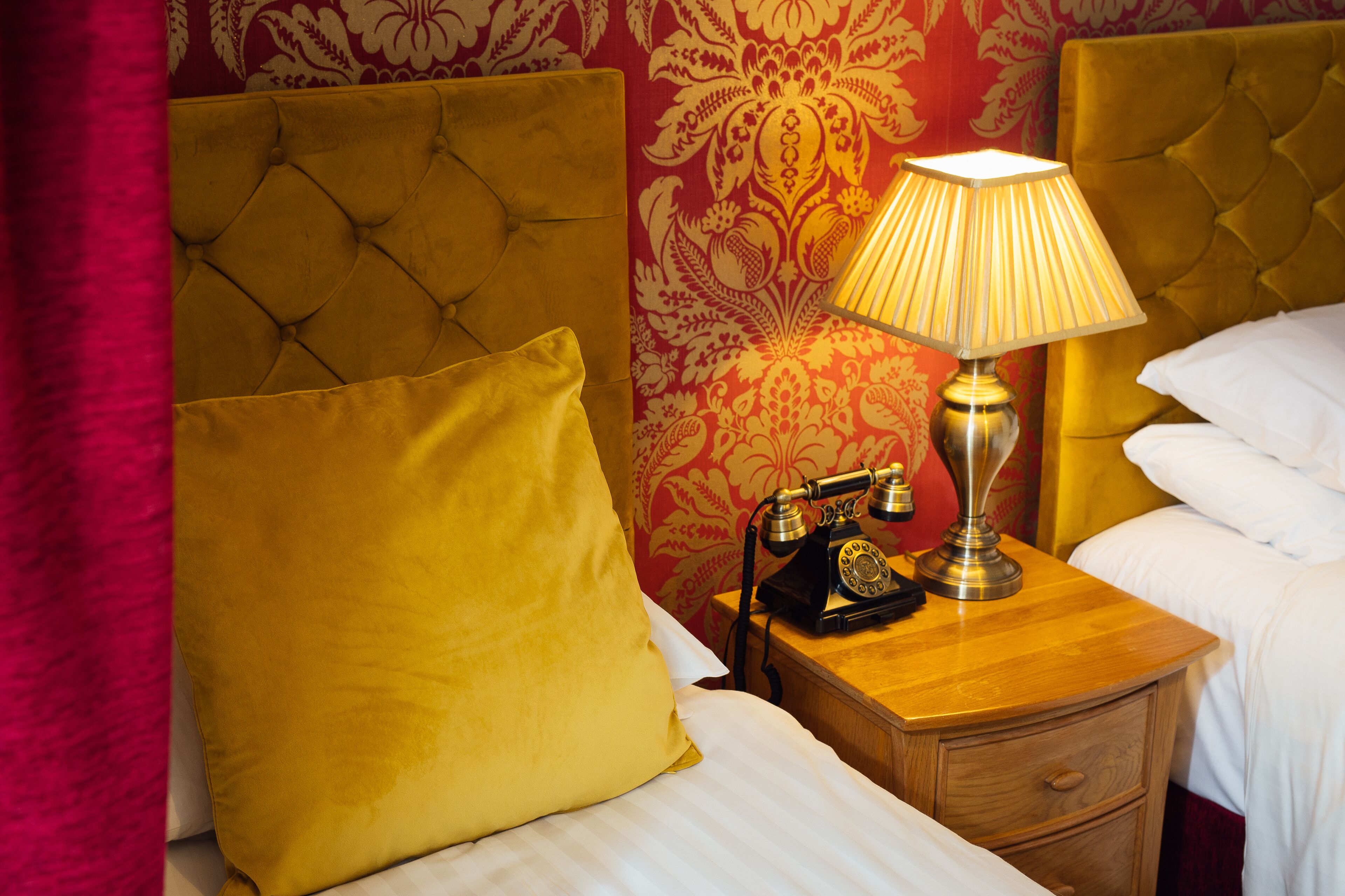 A hotel room with a tufted headboard, a wooden nightstand with a lamp and a rotary phone, a yellow pillow, and a white bed.