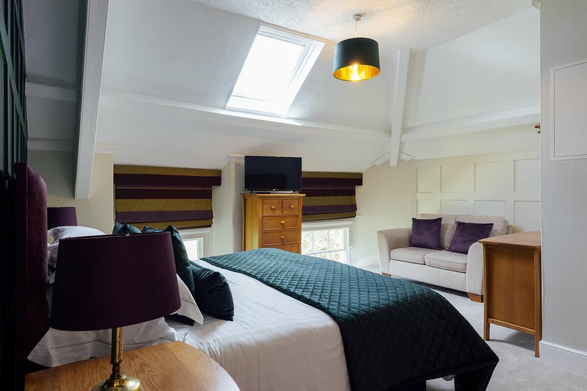 Une chambre à coucher comprenant un lit, un bureau, une commode, un téléviseur posé sur un meuble, un canapé et une fenêtre avec un puits de lumière.