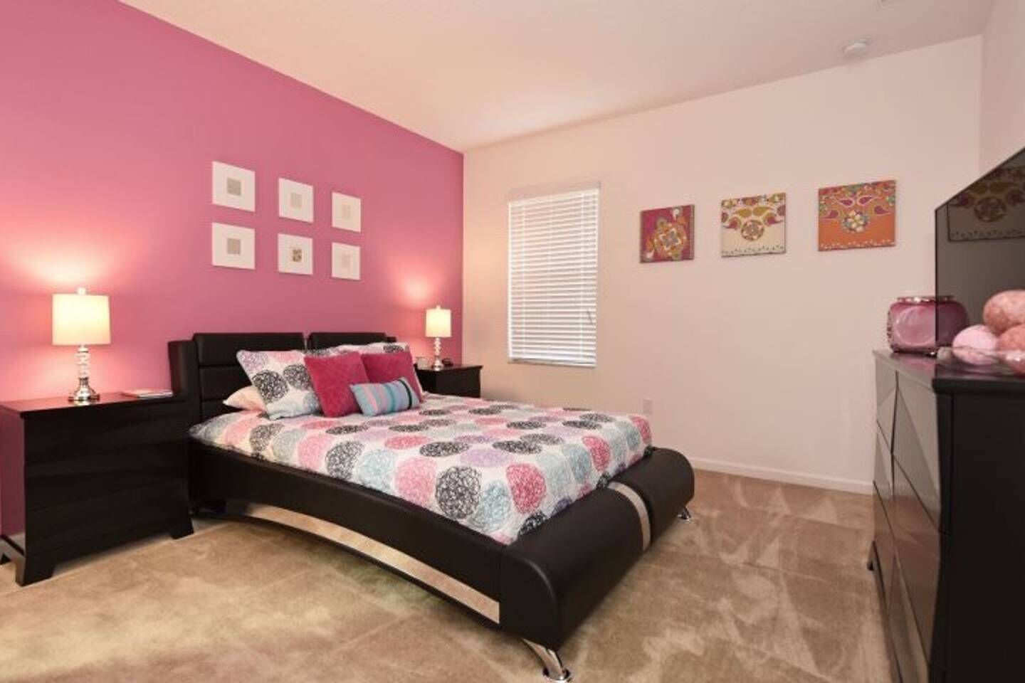 A bedroom with a pink wall, a bed with a patterned bedspread, two bedside tables with lamps, and a dresser with decorative items.
