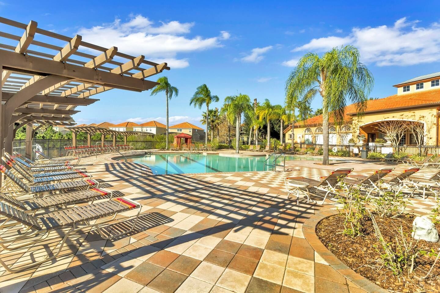 Zona de piscina con tumbonas y una pérgola, rodeada de palmeras y un cielo despejado.
