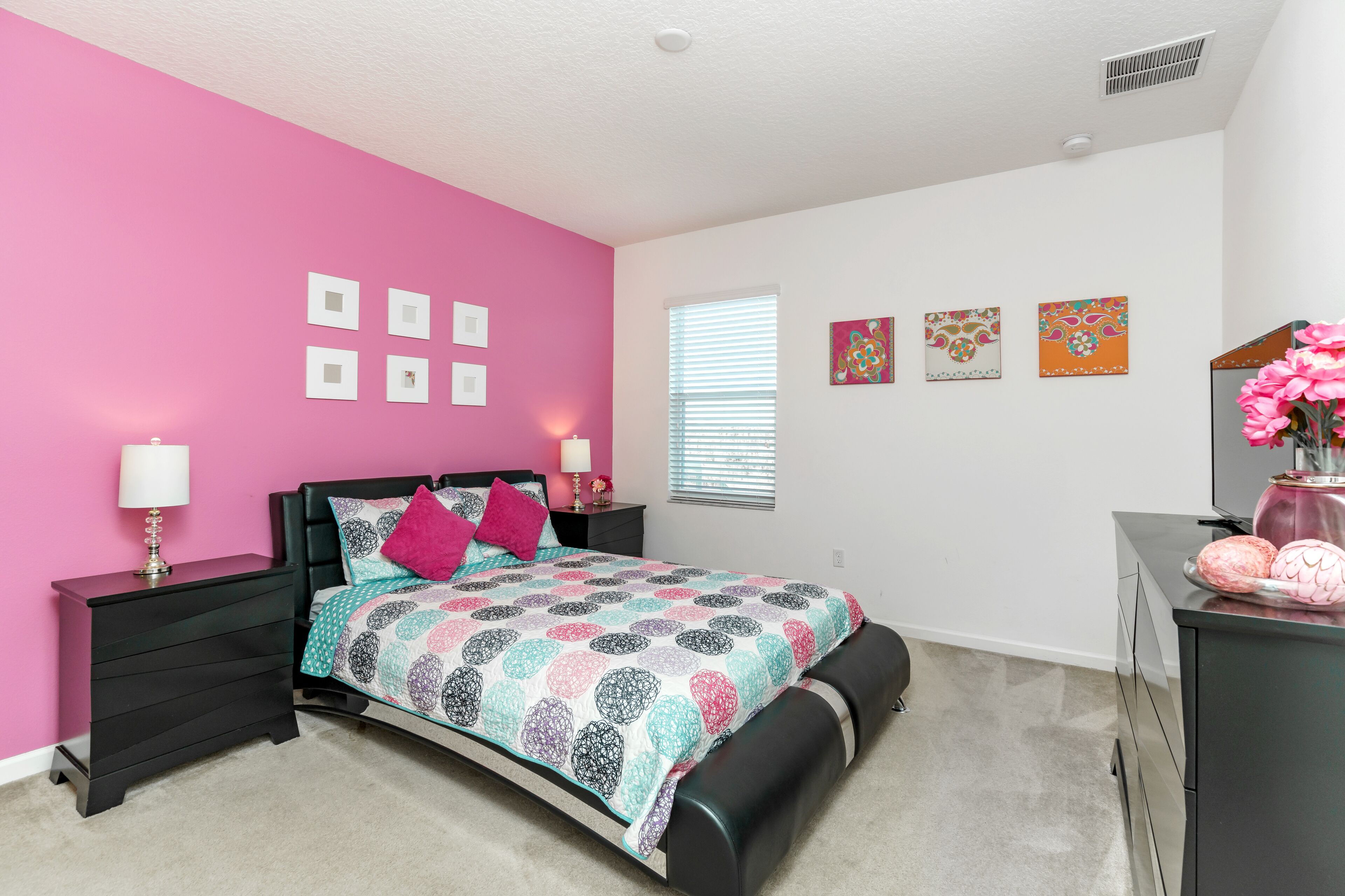 A bedroom with a pink wall, a bed with a patterned bedspread, a nightstand with a lamp, and a dresser with a mirror.