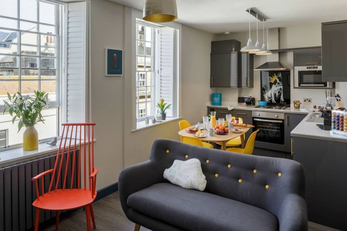 A modern kitchen with a dining area, featuring a grey sofa, red chairs, and a window with a view of buildings.