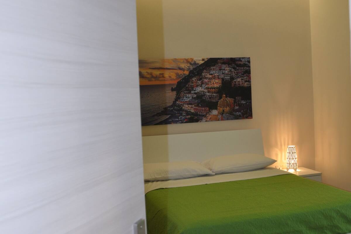 Quarto com cama, um quadro na parede, mesa de cabeceira com abajur e porta com adesivo.