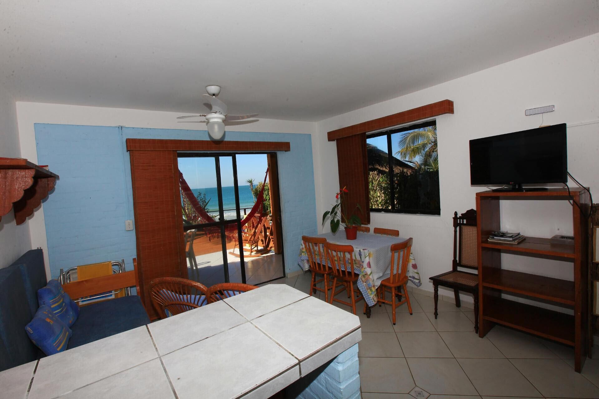 A living room with a TV, a ceiling fan, a dining area with a table and chairs, and a view of the ocean.